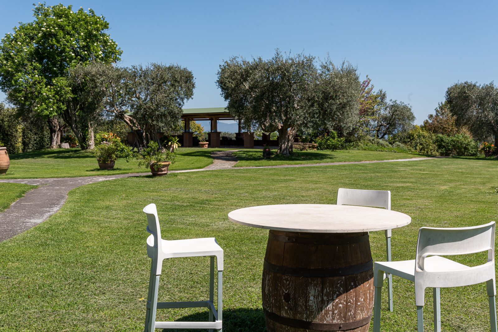 A round wooden table with two white chairs in a grassy outdoor area with trees and a building in the background.