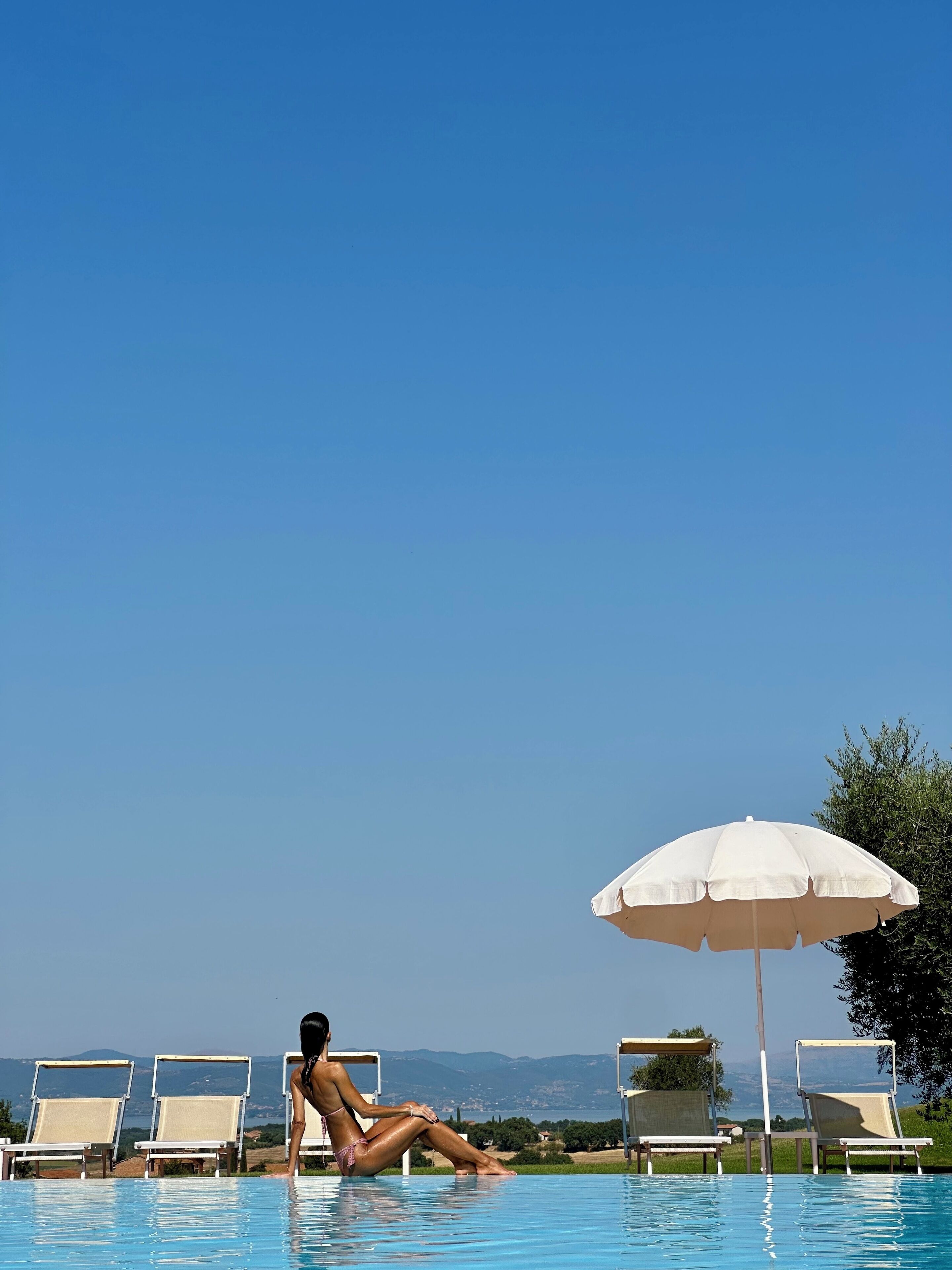 Blick auf ein Schwimmbecken mit einer Person, Liegestühlen und einem Sonnenschirm.
