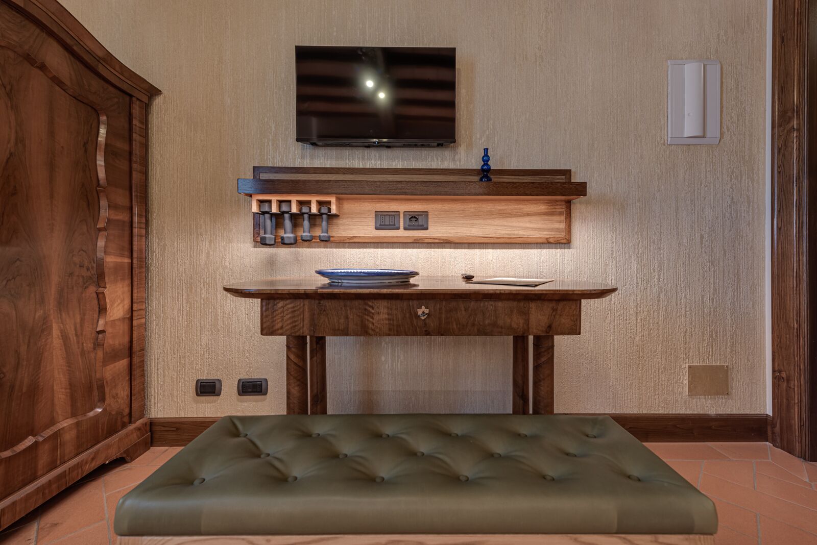 A room with a wooden cabinet, a television mounted on the wall, a wooden table with a bowl and a book, and a green cushioned bench.