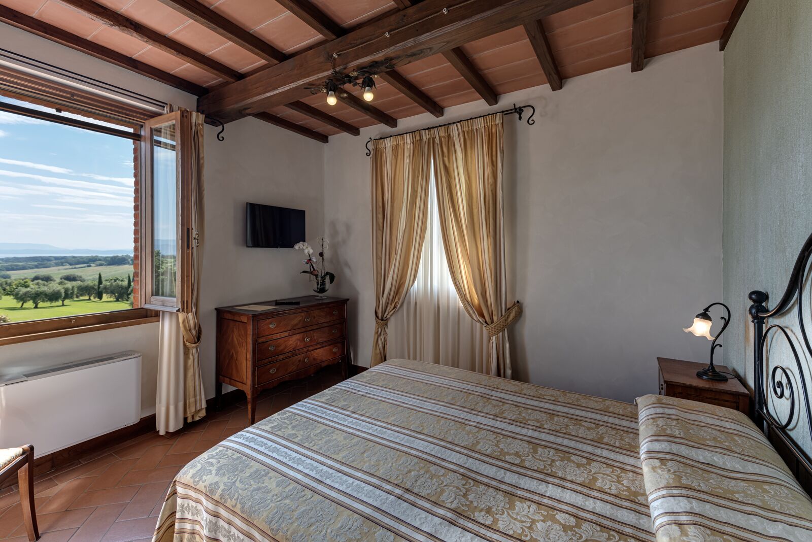 A bedroom with a bed, a dresser, a nightstand, a lamp, a TV, and a window with curtains.