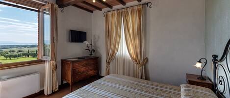 A bedroom with a bed, a dresser, a nightstand, a lamp, a TV, and a window with curtains.