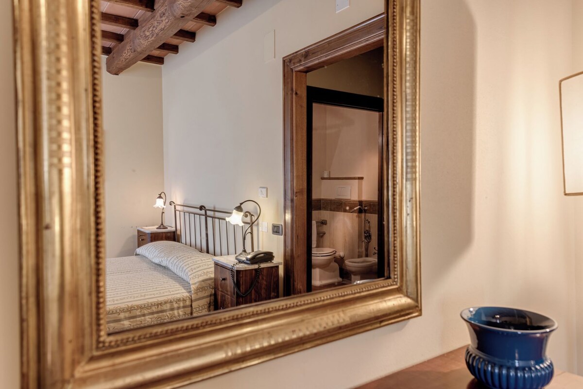 A bedroom with a bed, bedside lamps, a nightstand, and a mirror reflecting a bathroom with a toilet and shower.