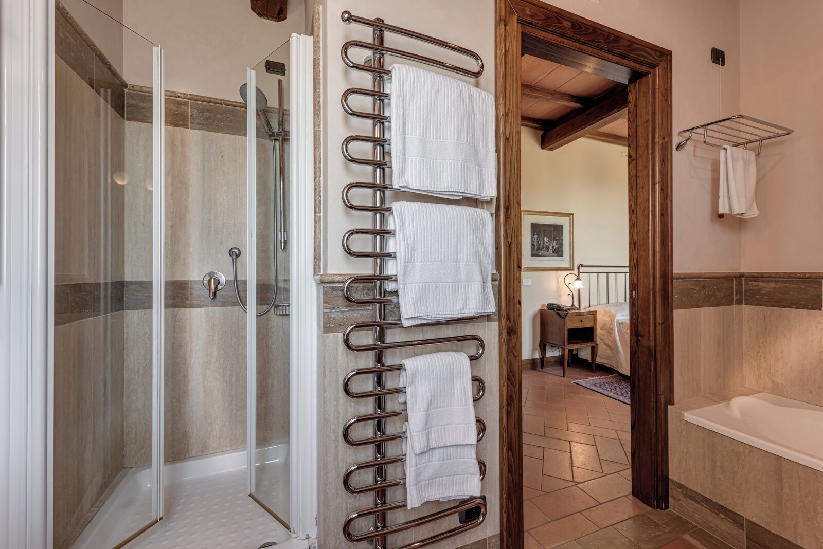 A bathroom with a glass shower enclosure, a metal towel rack with multiple towels, a wooden door frame, and a white bathtub.