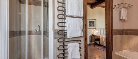 A bathroom with a glass shower enclosure, a metal towel rack with multiple towels, a wooden door frame, and a white bathtub.