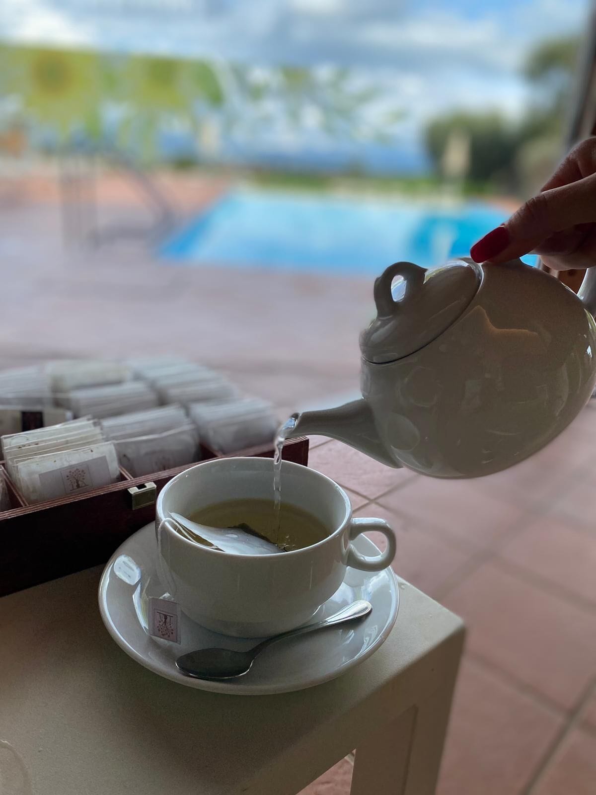 Una mano vertiendo té en una taza sobre una mesa con una bandeja de bolsitas de té y una cuchara.