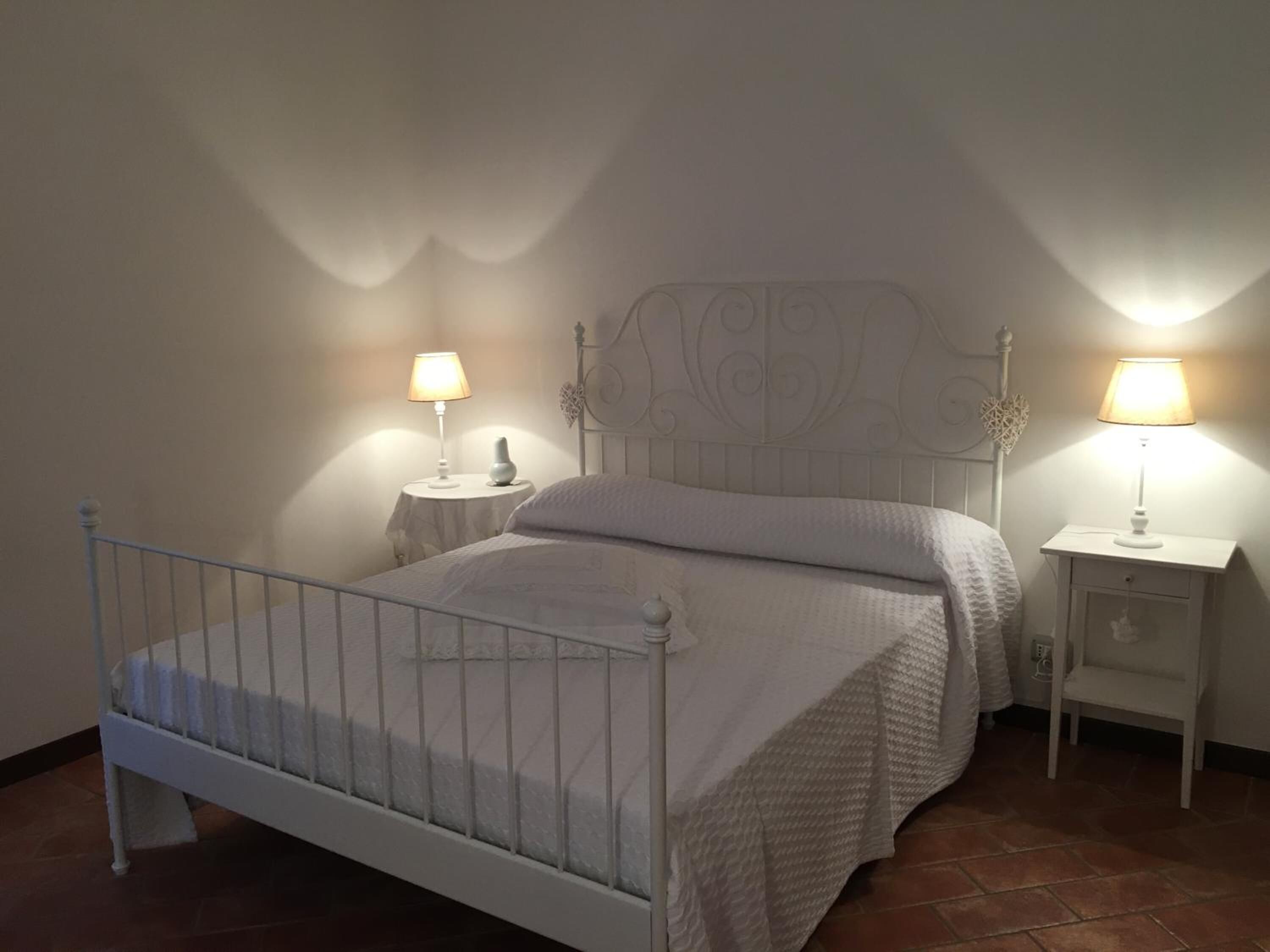 Ein Schlafzimmer mit weißem Metallbettgestell, einem weißen Kopfteil, zwei Nachttischen mit Lampen und einem kleinen Nachttisch mit einem dekorativen Gegenstand.
