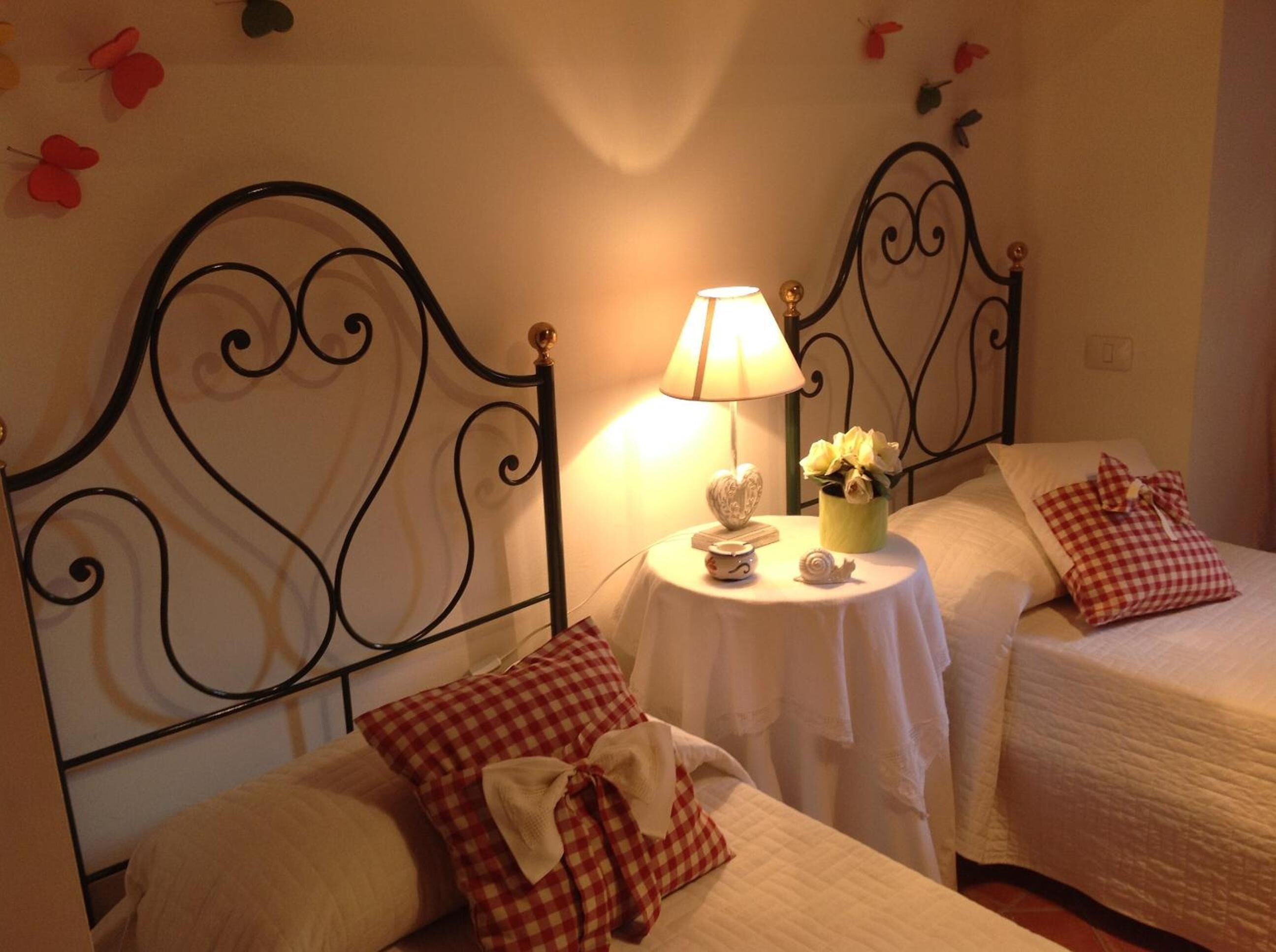 A bedroom with two single beds, each with a checkered pillow and a heart-shaped decoration. A bedside table holds a lamp, a small bowl, and a flower arrangement.
