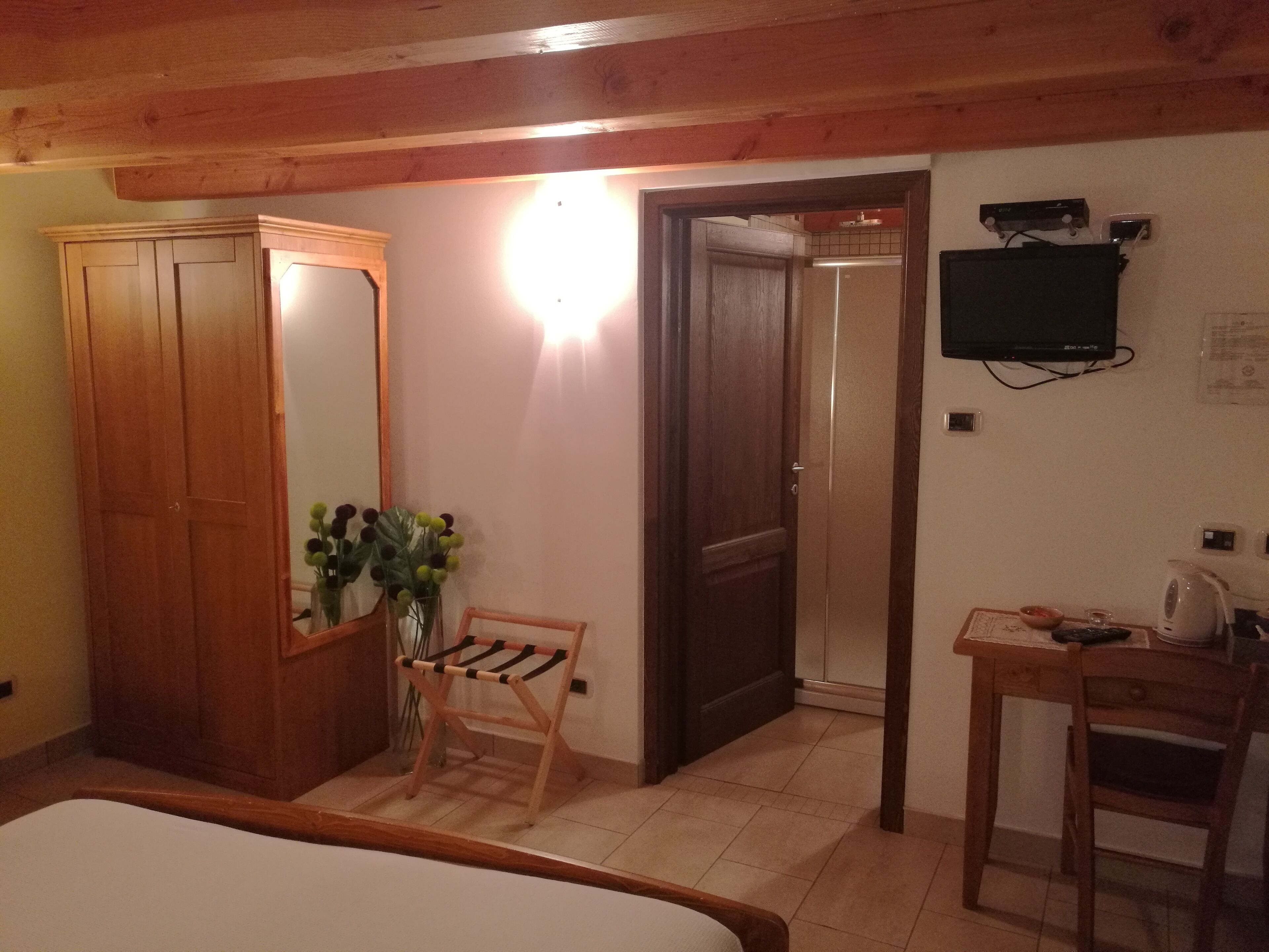 A bedroom with a wooden wardrobe, a bedside table with a kettle, a mirror, and a wall-mounted TV.
