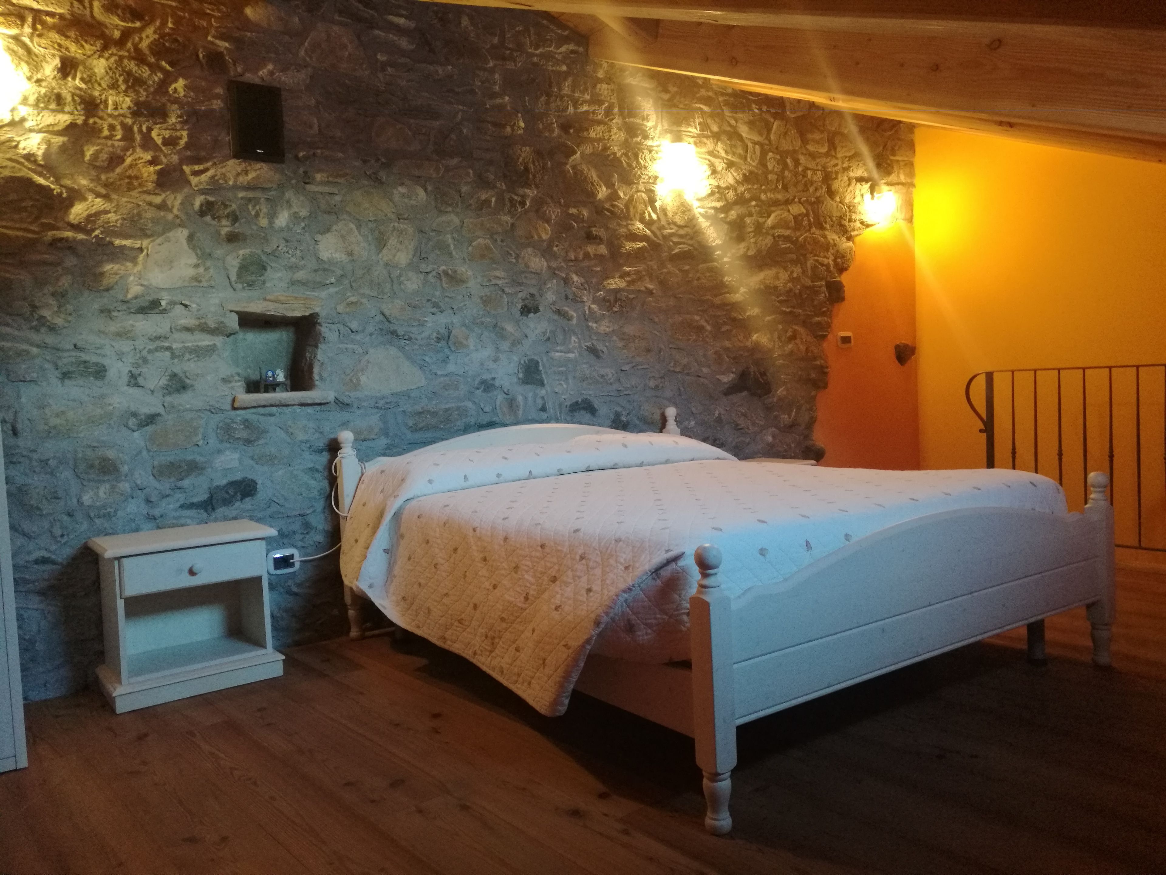 Un dormitorio con pared de piedra, cama de madera, mesita de noche y estante fijado a la pared.