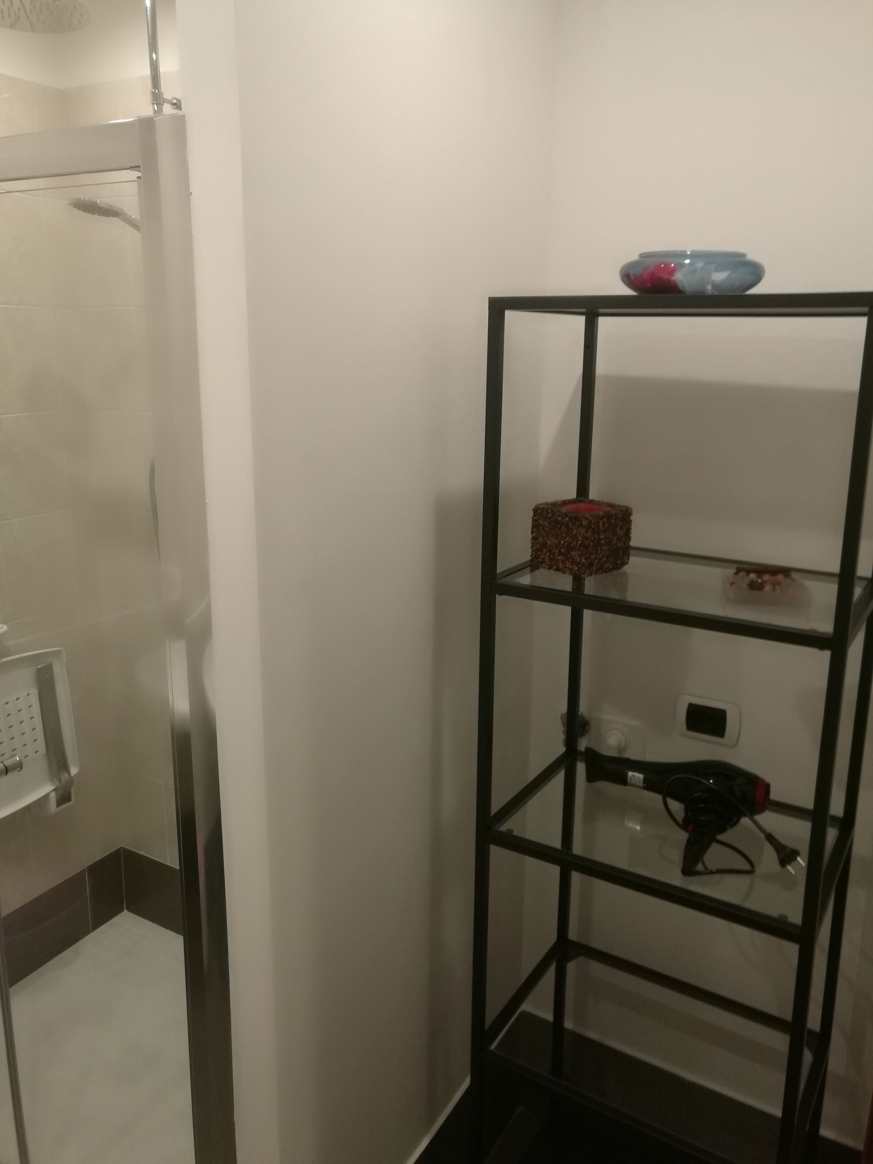 A bathroom with a glass shower enclosure, a black metal shelf unit with a red basket, and a hairdryer on the lowest shelf.
