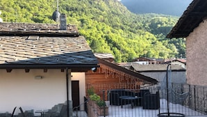 Un edificio rústico con un patio de piedra, mobiliario exterior y vistas a una ladera boscosa.