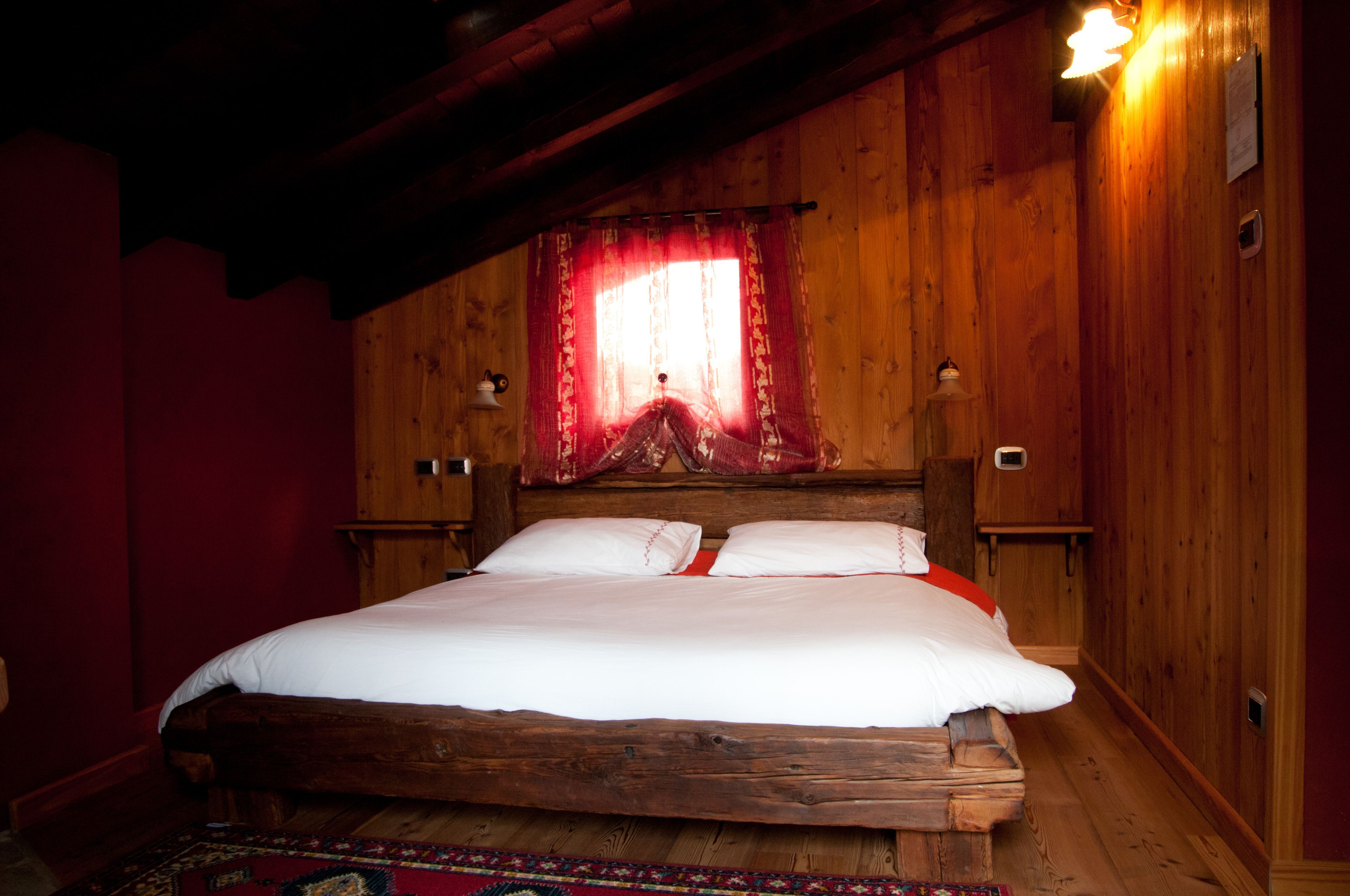A wooden four-poster bed with white linens, a red curtain, and two wall-mounted lamps.