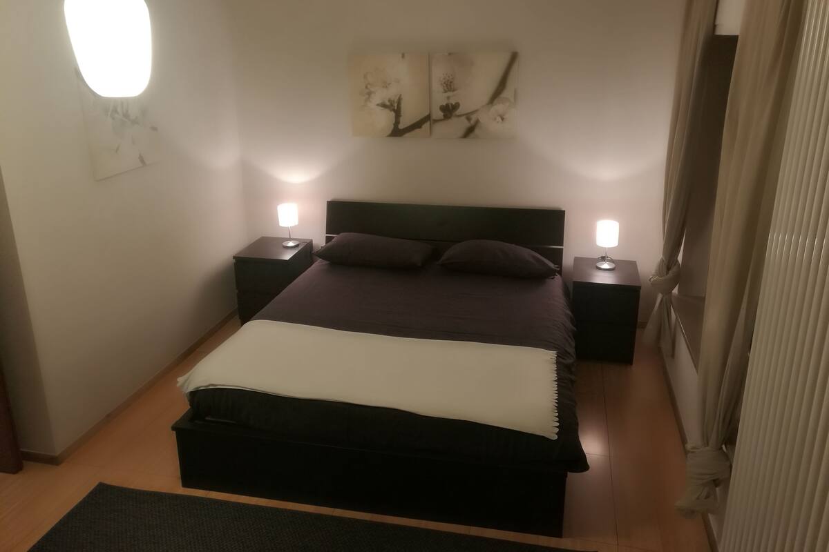 A bedroom with a large bed, two bedside tables with lamps, a wall art piece, and a window with curtains.