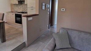 Una sala moderna con un sofá gris y un cojín escocés, una cocina con armarios de madera clara y salpicadero de azulejos, y una lámpara de techo.