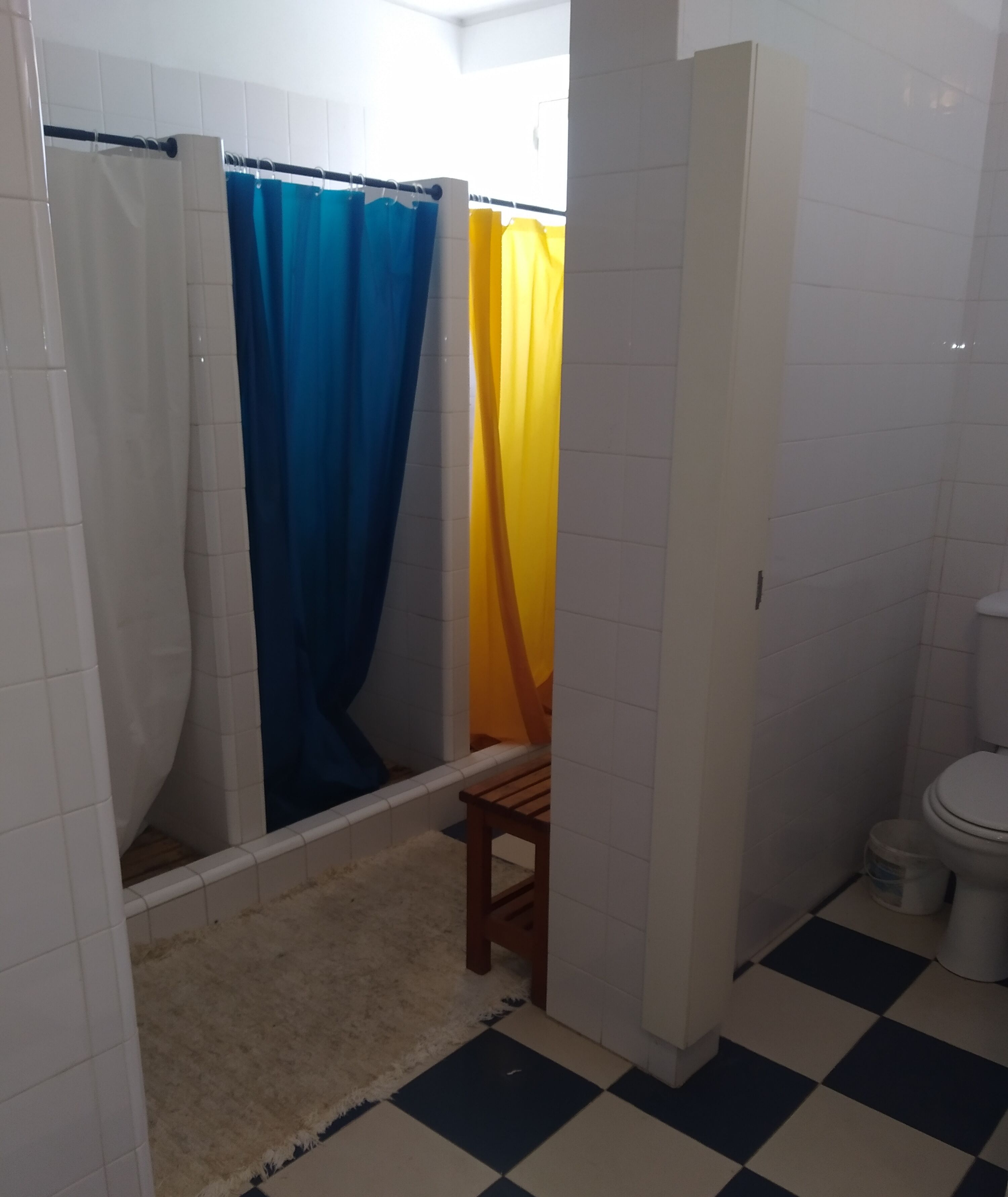 Un baño con piso a cuadros, un inodoro, una silla de madera y una ducha con cortinas azules y amarillas.
