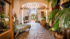 Un pasillo con piso de piedra, un banco acolchado, plantas en macetas y vista a un jardín exterior.