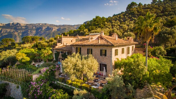 Una villa de piedra con techo de tejas, rodeada de exuberante vegetación y un telón de fondo montañoso.