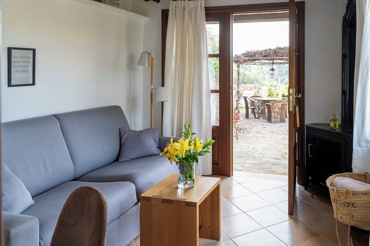 Ein Wohnzimmer mit grauem Sofa, einem Holztisch mit gelben Blumen, einer Lampe und Blick auf eine Außenpatio mit Stühlen und einem Tisch.