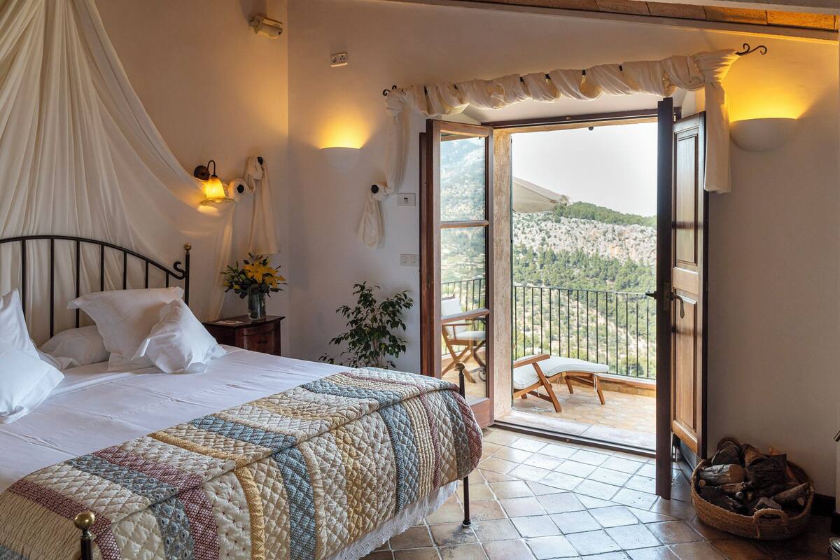 Ein Schlafzimmer mit einem Bett, einem Nachttisch mit Lampe, einer Zimmerpflanze und Blick auf einen Balkon mit Gartenmöbeln.