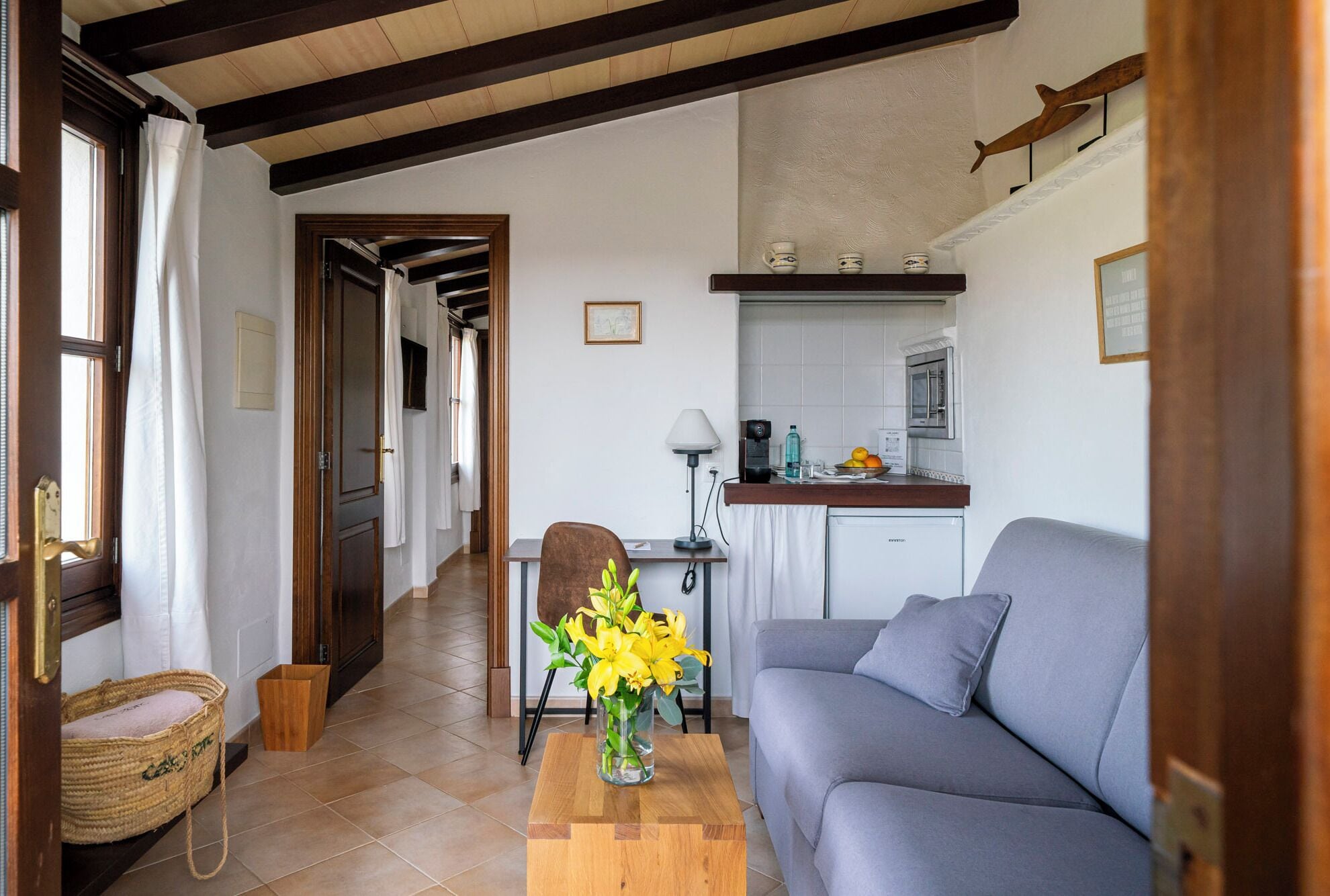 Una sala de estar acogedora con un sofá gris, una mesa de centro de madera con un jarrón de flores amarillas, y una zona de cocina con un fregadero, estufa y encimera.
