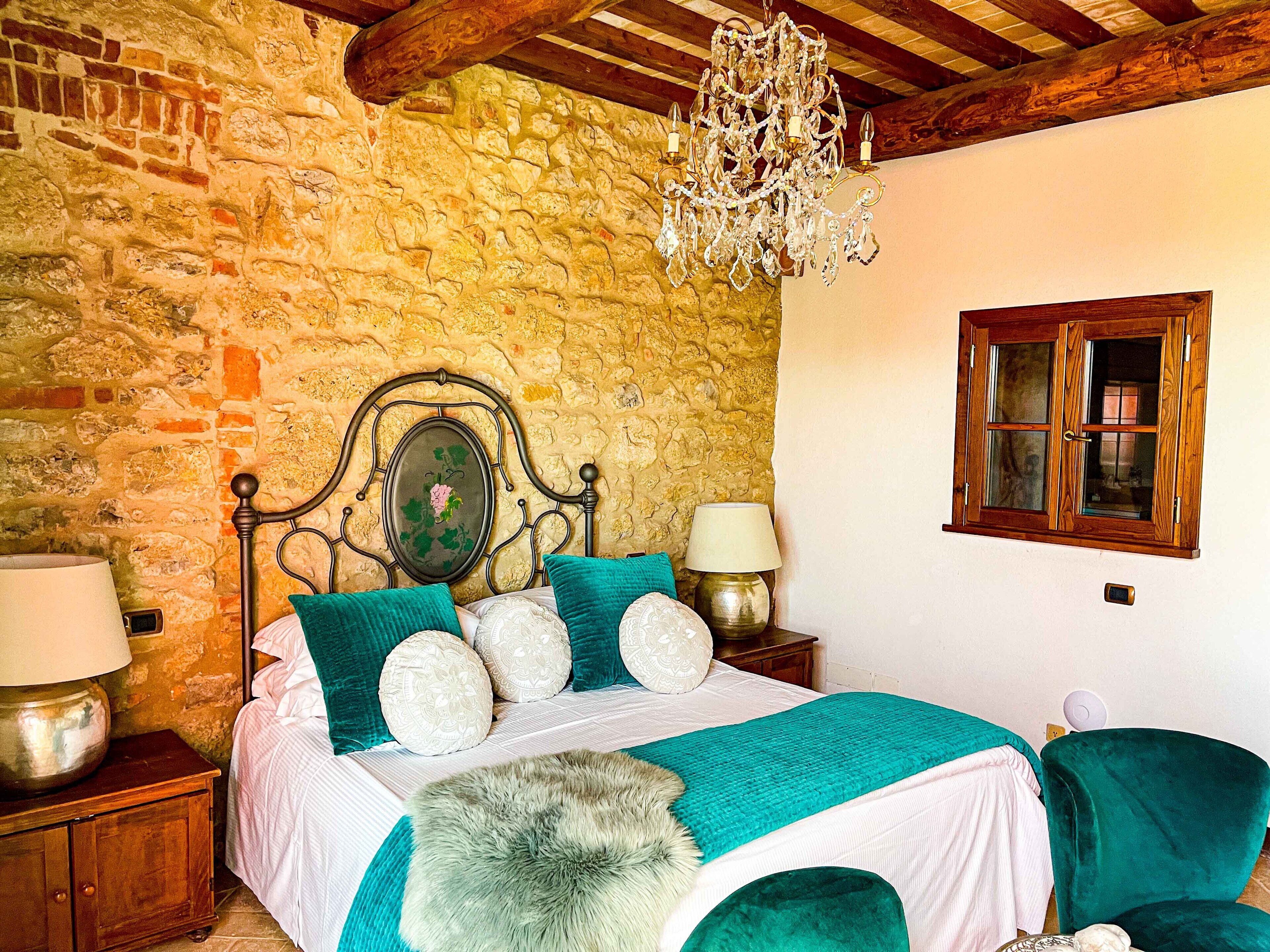 A bedroom with a stone wall, wooden ceiling beams, a chandelier, a bed with teal bedding, and a window with wooden shutters.