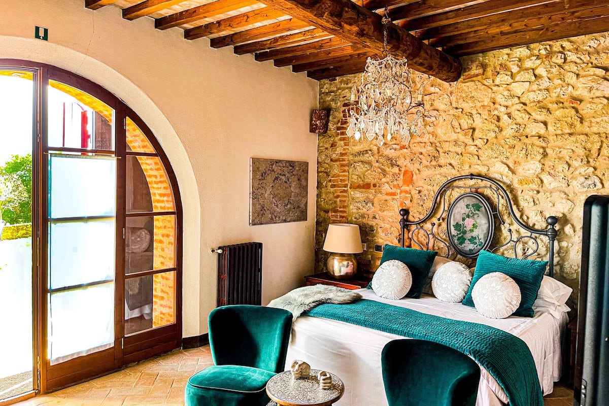 A bedroom with a stone wall, a bed with teal bedding, a chandelier, and a view through a glass door.