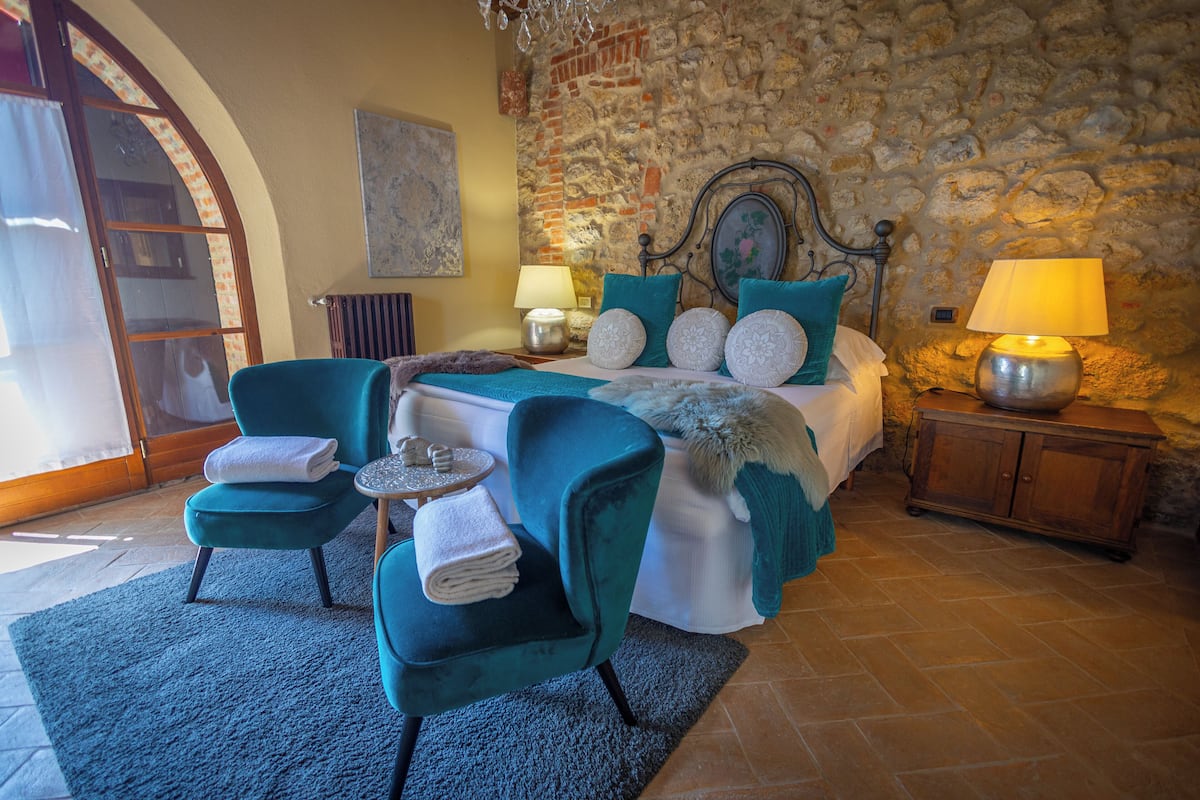 A bedroom with a stone wall, a bed with teal bedding, two chairs, a nightstand with lamps, and a wooden cabinet.