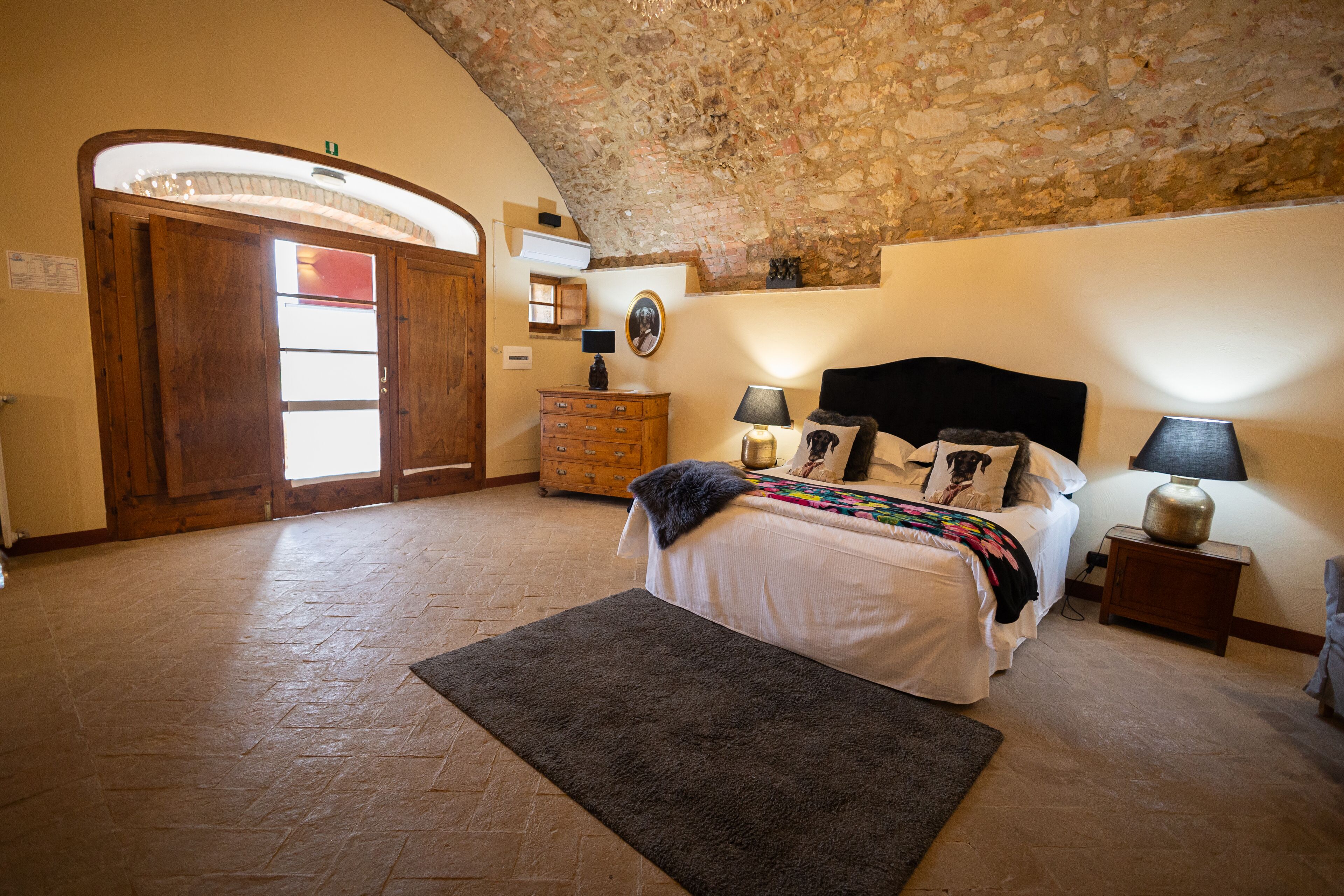 A bedroom with a stone ceiling, a bed with a black headboard, two bedside tables with lamps, a wooden dresser, and a door with a glass panel.