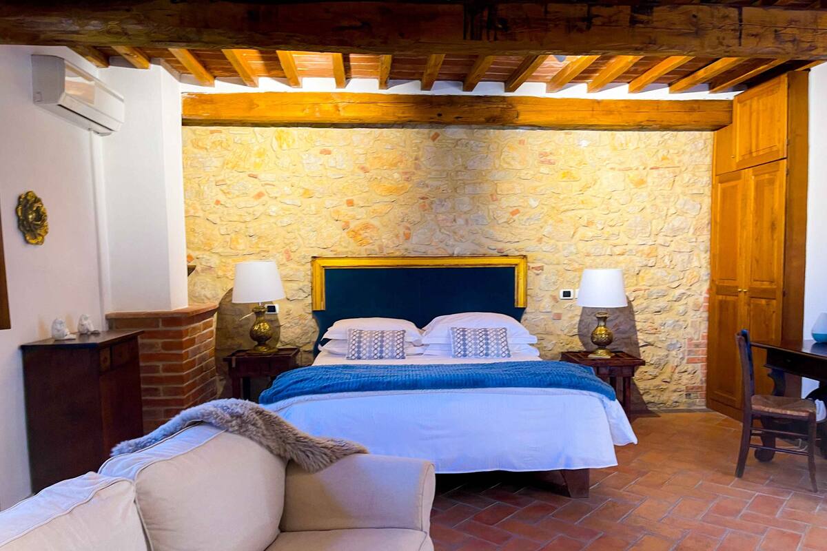 A bedroom with a stone wall, wooden ceiling beams, a bed with two pillows, a nightstand with a lamp, and a wooden chair.