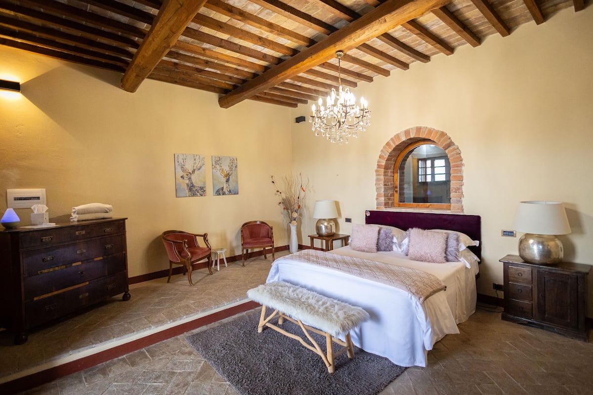 A bedroom with a large bed, a bench, a dresser, a chair, a lamp, and a chandelier.