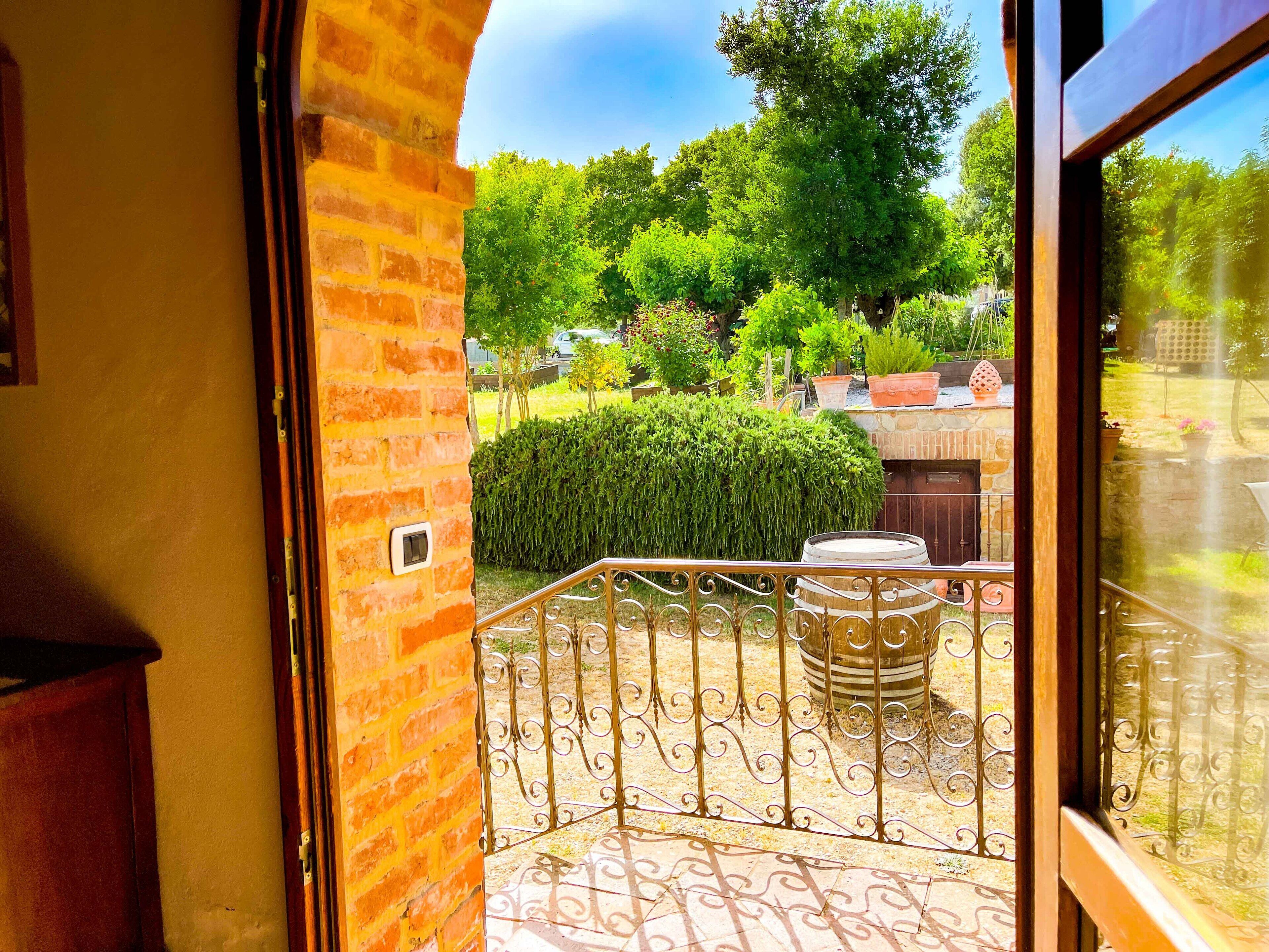 Un balcón con barandilla de hierro forjado, un barril y vista a un jardín con árboles y césped.