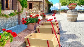Un edificio rústico de piedra con tejado de tejas, contraventanas de madera y un patio de grava. Se ven carretillas de madera y macetas con flores rojas.