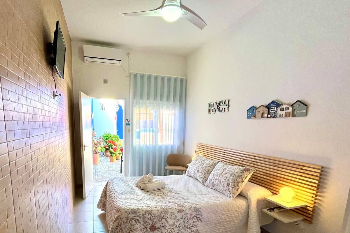 A bedroom with a bed, a bedside table with a lamp, a ceiling fan, and a view of the outside.