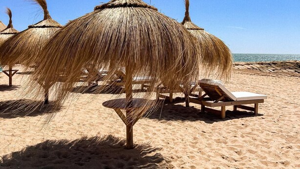 Una playa con sombrillas de paja y sillones de madera.