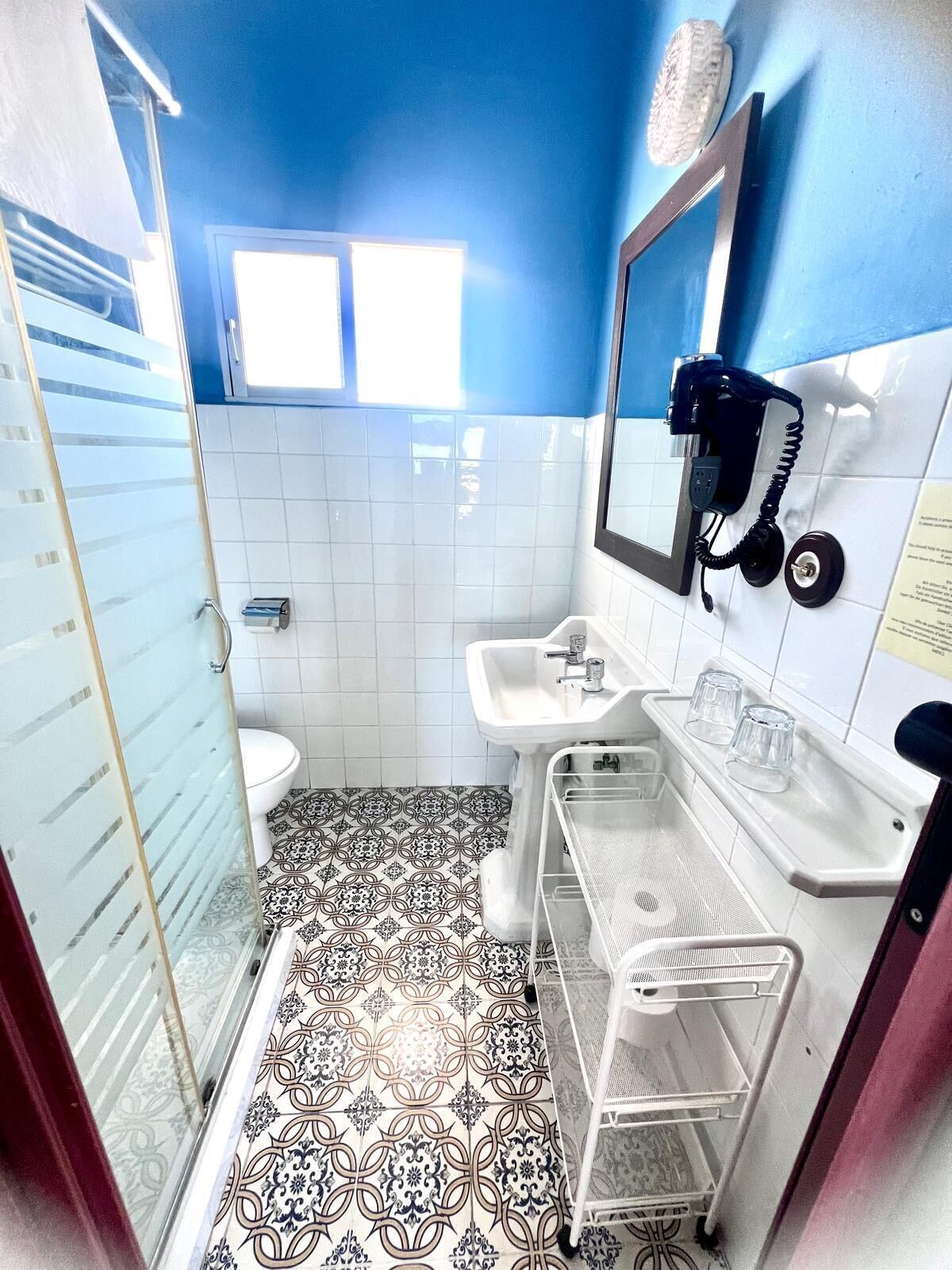 A bathroom with a toilet, sink, and a mirror. There is a towel rack and a shelf with glasses. The floor has patterned tiles.