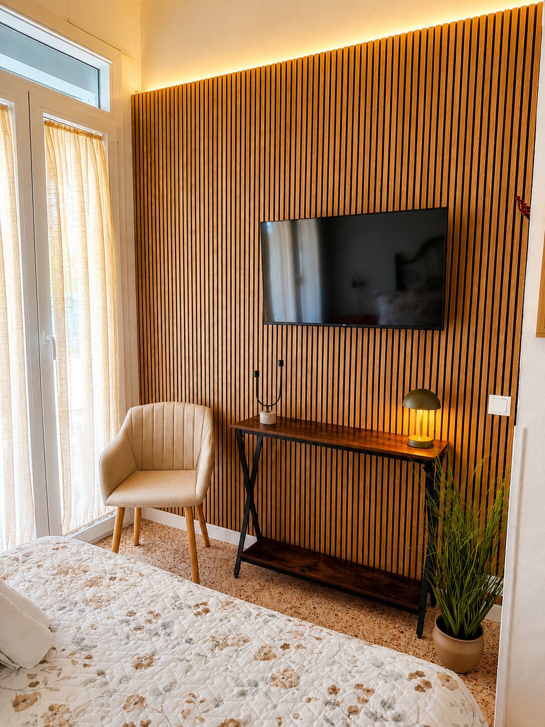 A modern room with a TV mounted on a wooden panel wall, a sofa, a bedside table with a lamp, and a potted plant.