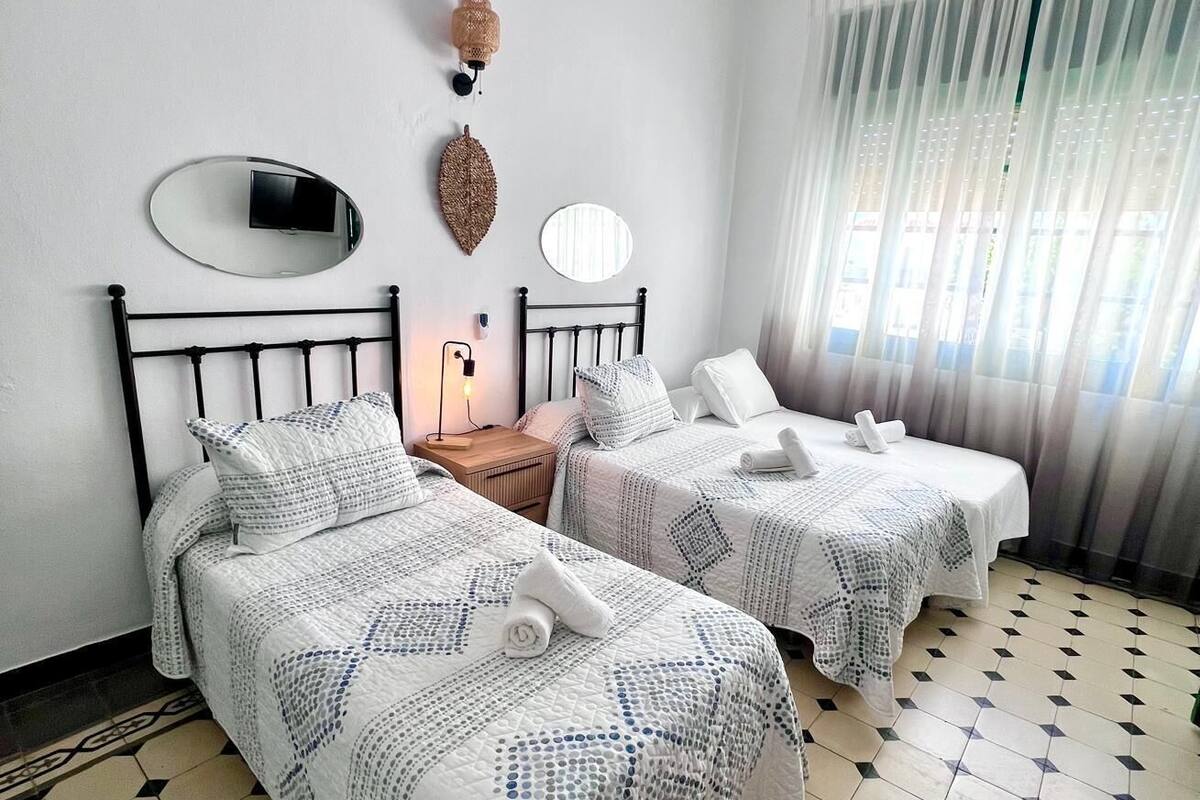 A hotel room with two beds, a mirror, a wall-mounted lamp, and a window with sheer curtains.