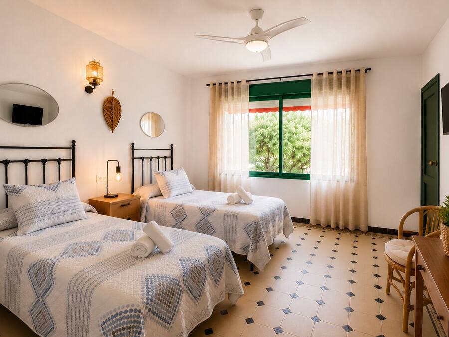 A hotel room with two beds, a ceiling fan, a television, a window with curtains, and a dining area with a chair and table.