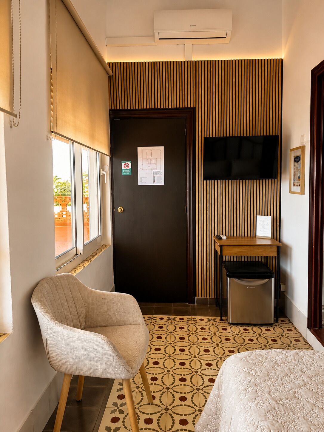 A hotel room with a bed, a chair, a desk, a trash bin, a TV, and a door with a sign.