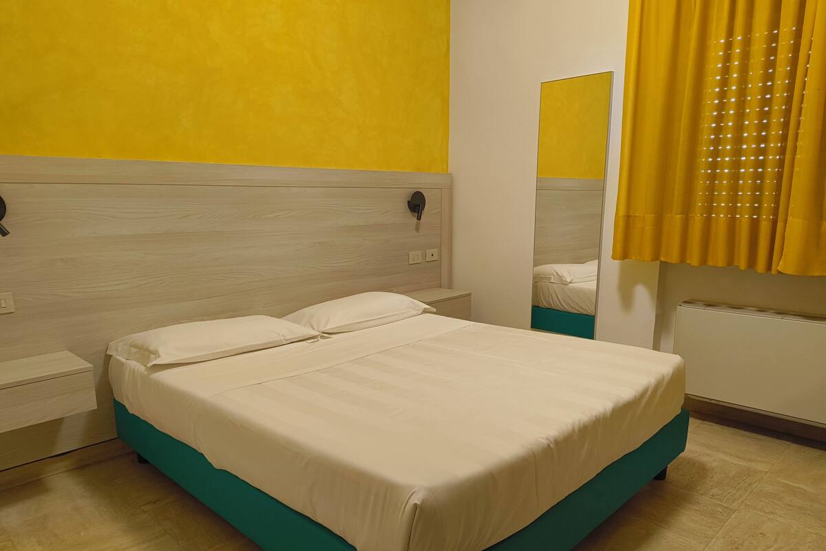 A bed with a teal frame and white bedding in a room with yellow walls and a mirror.