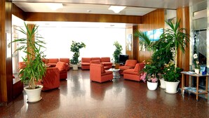 Een ruime lobby met een gepolijste vloer, rode banken en diverse potplanten.