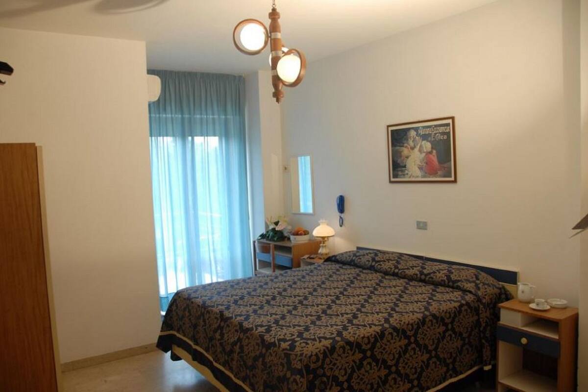 Un dormitorio con cama, una mesita de noche con lámpara, un cuadro enmarcado en la pared y una ventana con cortinas.