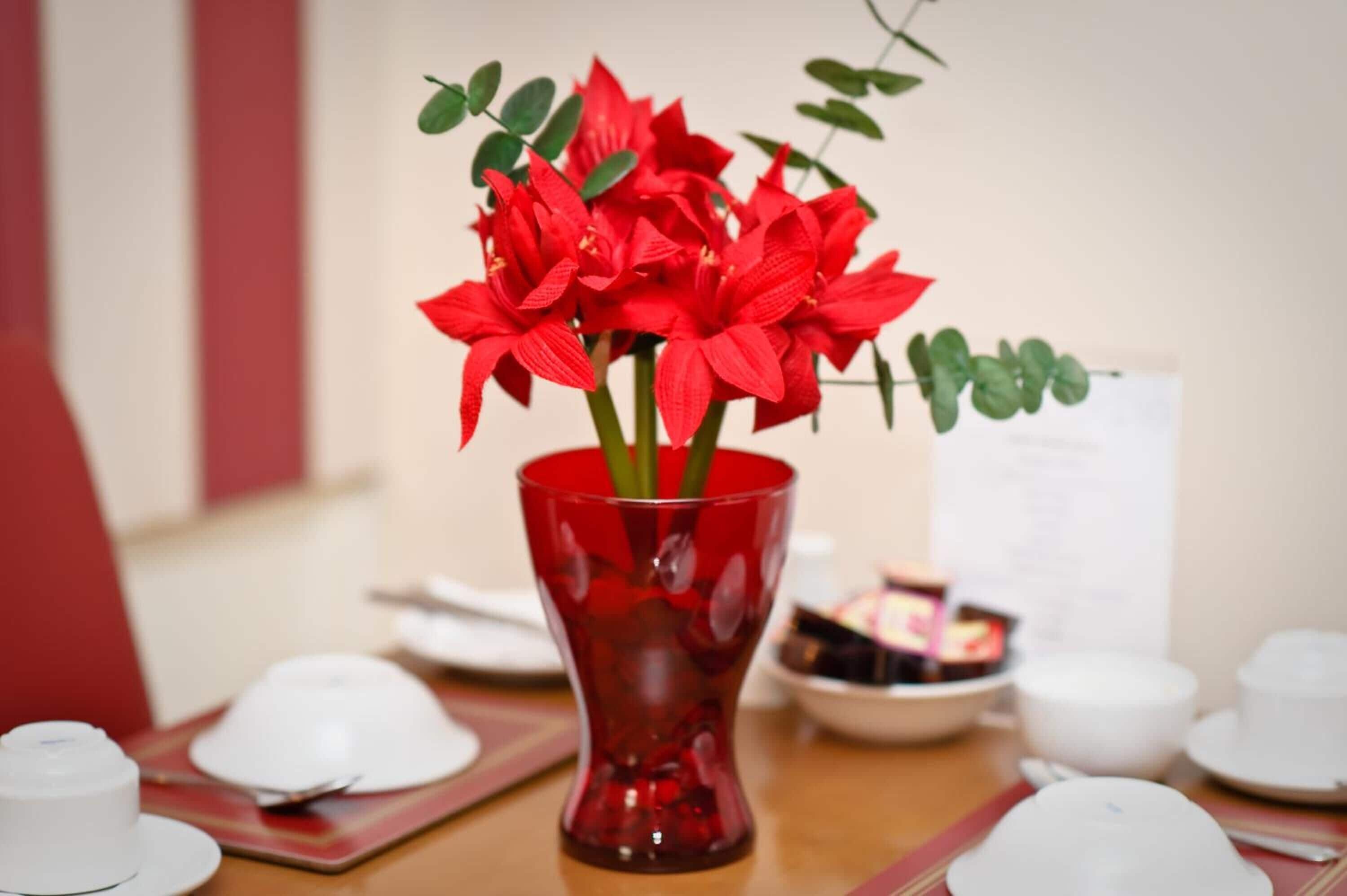 A table with a red vase containing red flowers, white teacups, and a menu.
