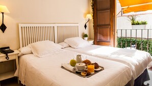 Una cama bien hecha con sábanas blancas, una bandeja con alimentos para el desayuno, incluyendo un croissant, un muffin y jugo de naranja, y una mesita de noche con una lámpara y un teléfono.