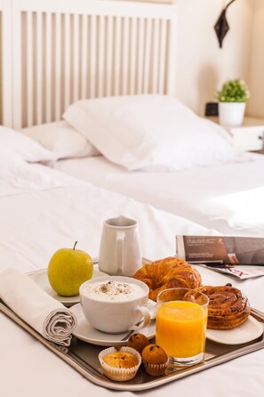 A breakfast tray with a croissant, muffin, apple, coffee cup, and orange juice on a bed with white bedding and a headboard. - Miguel de Cervantes (Alcala de Henares)