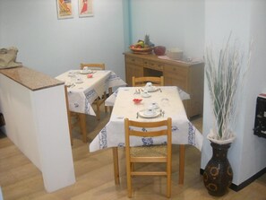 A dining area with two tables set for four, each with white tablecloths and blue floral patterns. There are wooden chairs, a small cabinet with a fruit bowl, and a vase with tall, thin plants. - Hotel Colombo (Riccione)