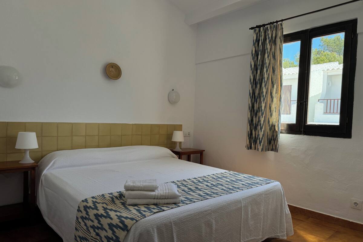 Una habitación de hotel con cama, dos mesitas de noche, una ventana con cortinas y vistas al exterior.