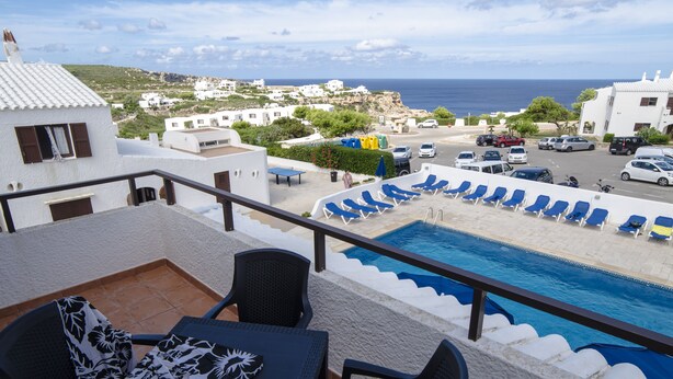 A poolside area with lounge chairs, a table, and a view of the sea and buildings in the background. - Apartamentos Sa Cala (Ciutadella de Menorca)