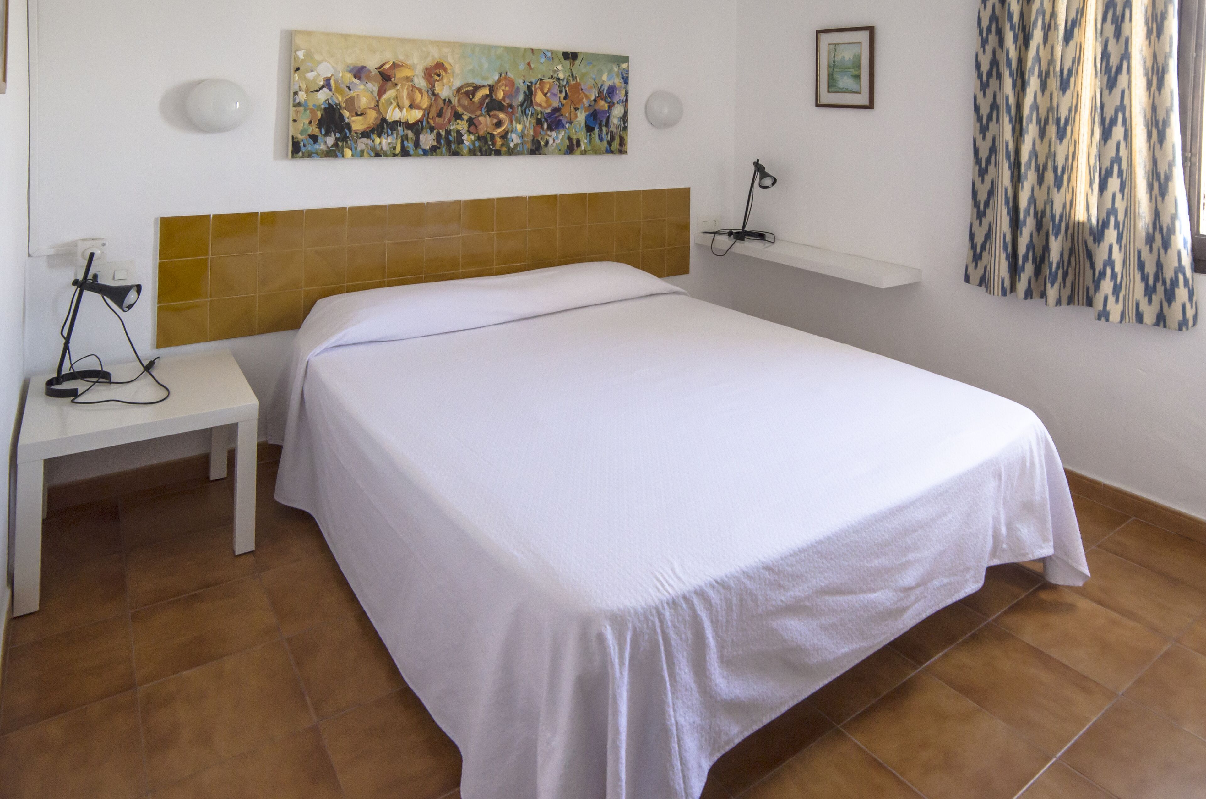 Une chambre d’hôtel avec un lit, une table de chevet, une lampe, un tableau et une fenêtre avec des rideaux.
