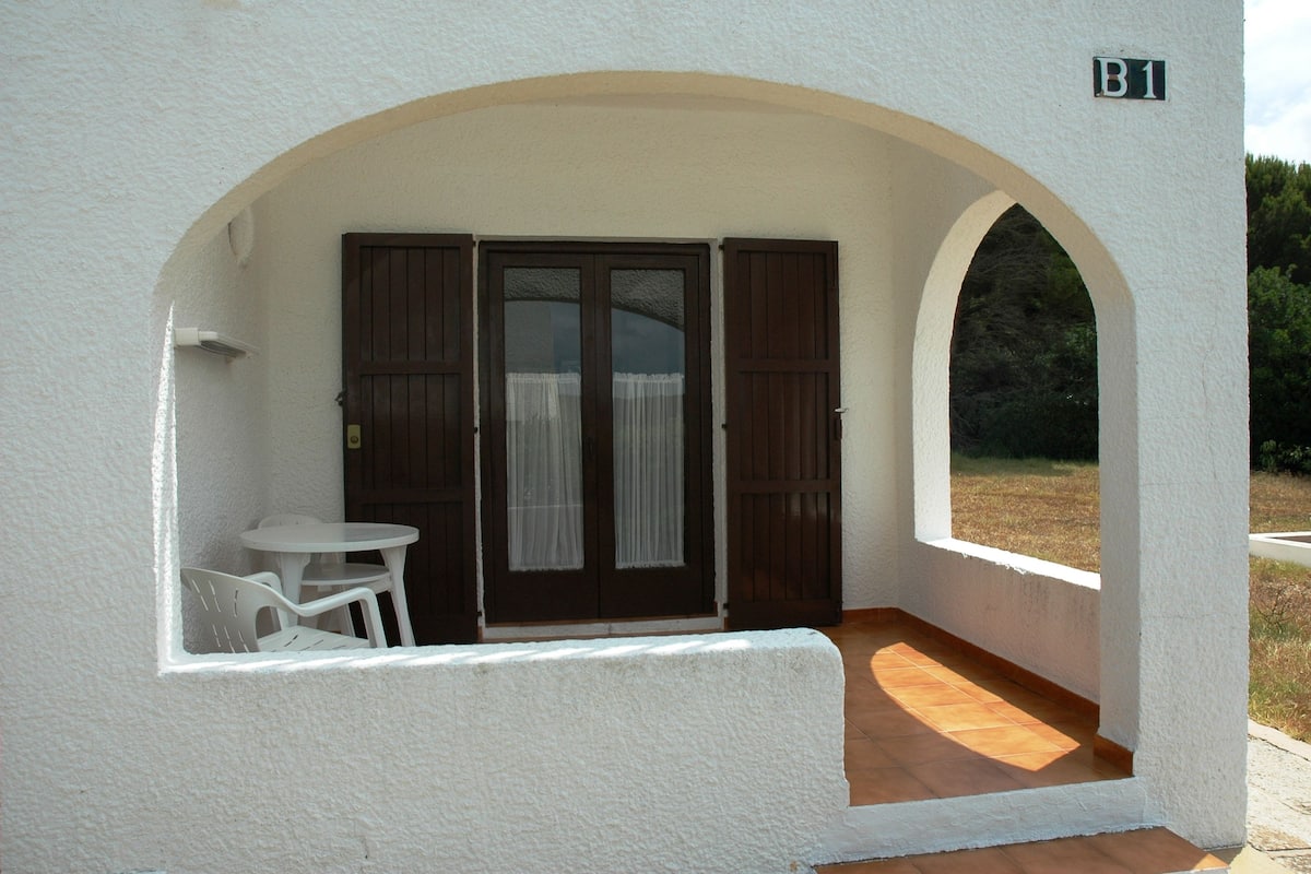 Un edificio blanco con un patio techado, una puerta con cortinas y una pequeña mesa con sillas.